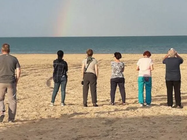 Group of people standing on a sandy beach looking at a rainbow over water