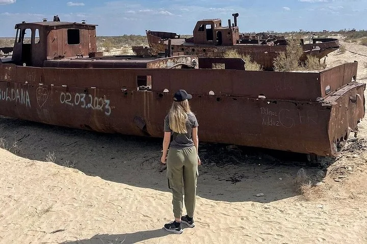 A woman looks at a rusted ship covered in graffiti in the desert.