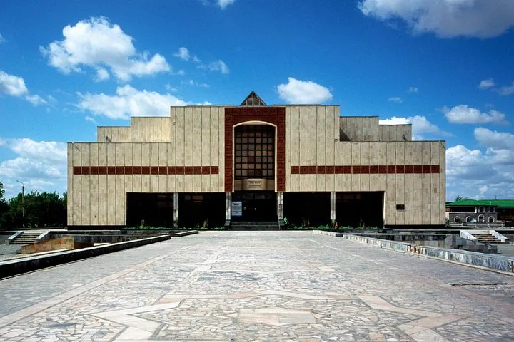 A large, modern, light-colored building with a wide paved courtyard under a blue sky.