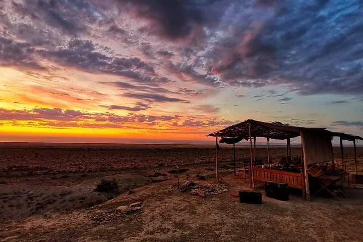 A wooden shelter stands on a hill overlooking a vast plain during a colorful sunset.
