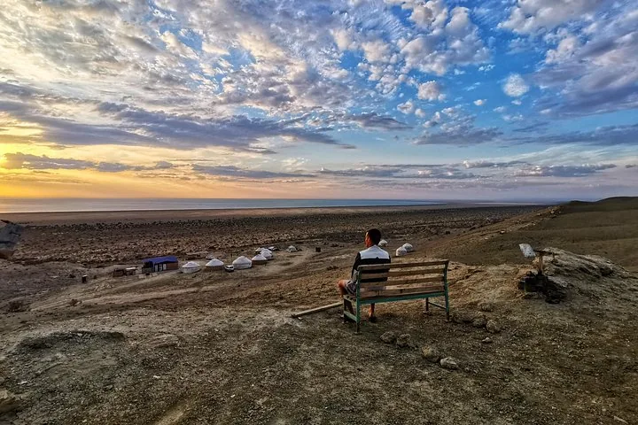 A person sits on a wooden bench overlooking a yurt camp and the Aral Sea.
